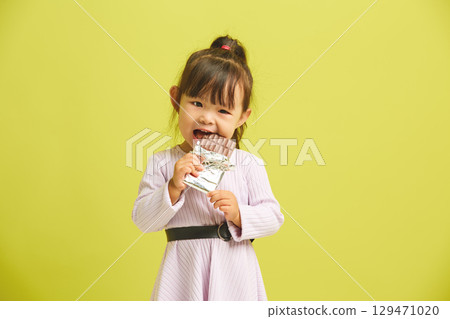 Small female child having addiction to sugar eats bar of chocolate standing over yellow isolated background. Sweet tooth concept. 129471020