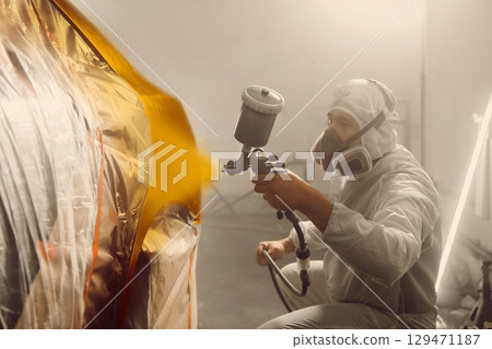 Auto mechanic worker spraying car body to orange gold color using spray gun in a paint chamber during repair work. 129471187
