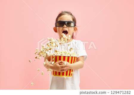 Joyful and Surprised Child Girl in 3D Glasses Holding Bucket of Popcorn Which is Flying to Sides Standing Over Pink Isolated Background. 3D Movie Thrill 129471282