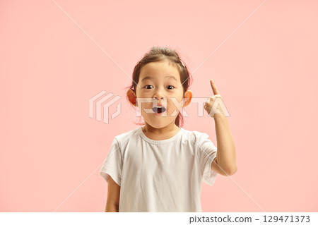 Little Girl with a Surprised Expression Points Up Above her on Free Copy Space, Expresses Joyful and Excited, Showing With Finger Upwards and Looking To Camera with Wide Open Eyes and Mouth 129471373
