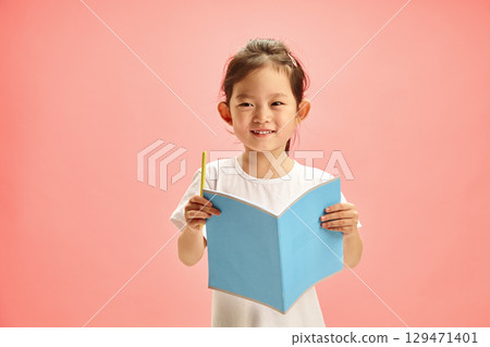 Happy and Pretty Preschool-age Korean Girl with an open Notebook with Cover Facing out and Holding Pen, Smiling Sweetly Standing on a Pink Isolated Background. Concept of Remote Learning. Happy and Pretty Preschool-age Korean Girl with an open Notebook with Cover Facing out and Holding Pen, Smiling Sweetly Standing on a Pink Isolated Background. Concept of Remote Learning. 129471401