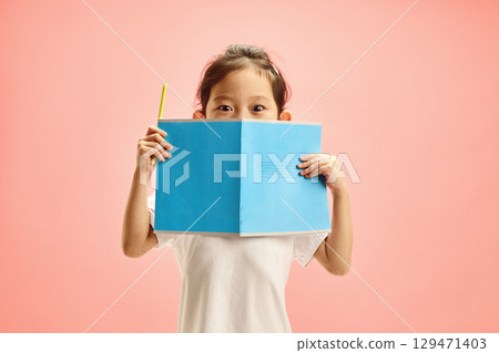 Curious Elementary Age Child Girl with Big Eyes Looks Out from Behind an open Blue Notebook with Cover on Outside, Expresses Surprised and Interest in Knowledge Standing Over Pink Isolated Background 129471403