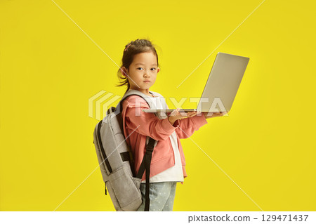 Little Asian Kid with Laptop and School Backpack stands over yellow isolated background 129471437