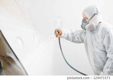 Male repairman painter in chamber painting automobile car elements. Worker paints the vehicle in white with a spray gun. 129471571