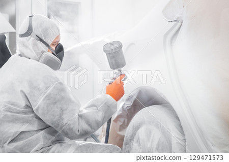 Body painter with airgun spraying white paint on a automobile in car maintenance service paint room. 129471573