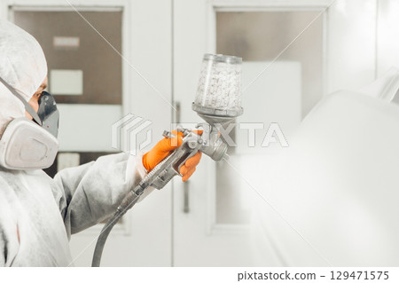 Male worker sprays paint with spray gun on car body part in car maintenance service paint room. Male worker sprays paint with spray gun on car body part in car maintenance service paint room. 129471575