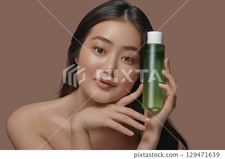 Beautiful young asian woman with a clean smooth skin holding green tube of facial tonic near her face posing on a beige background with a free copy space. Beautiful young asian woman with a clean smooth skin holding green tube of facial tonic near her face posing on a beige background with a free copy space. 129471639