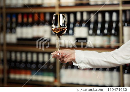 Glass of wine filled with white wine in female hand on cellar and bottles shelf background, close up with a free copy space for your text.  129471680