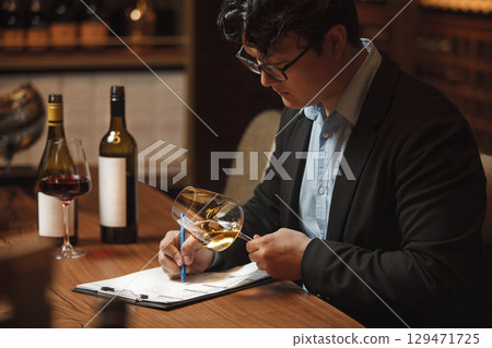 Wine Expert Evaluates Color of White Wine Over Clean Paper Background Sitting at Table in Cellar 129471725