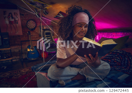 Teen girl reading book in cozy attic with guitar and retro decor 129472445