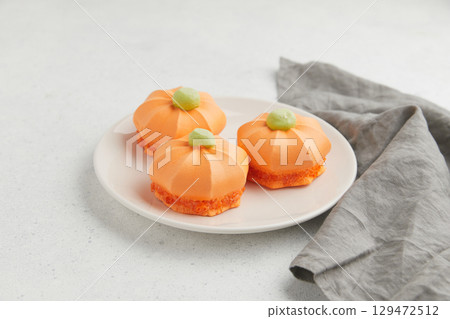 Three orange pumpkin shaped desserts with green tops, placed on a white plate with a gray cloth nearby 129472512