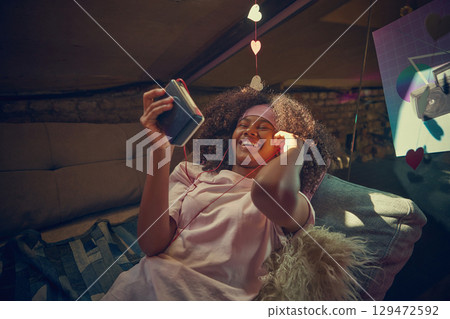 Smiling teenage girl relaxes in cozy attic, listening to vintage cassette player with headphones. 129472592