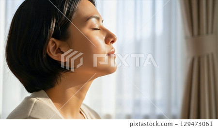 Peaceful Asian woman enjoying sunlight by window in serene morning moment 129473061