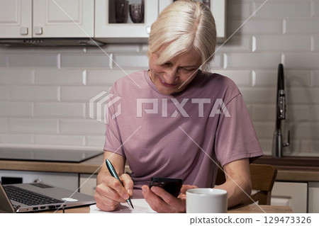 Focused Senior Woman Engaged in Multi-tasking with Phone and Notepad at Kitchen Table, Balancing Work and Daily Life Activities Focused Senior Woman Engaged in Multi-tasking with Phone and Notepad at Kitchen Table, Balancing Work and Daily Life Activities 129473326