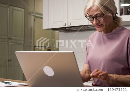 A Joyful Older Woman Engaging with Technology at Home, Demonstrating Digital Skills While Using a Laptop in a Cozy Kitchen Setting A Joyful Older Woman Engaging with Technology at Home, Demonstrating Digital Skills While Using a Laptop in a Cozy Kitchen Setting 129473331