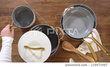 A person lifts a grey pot from a neatly arranged cookware set on a wooden kitchen surface. White lids with gold handles and wooden spoons are visible in the layout. 129473487