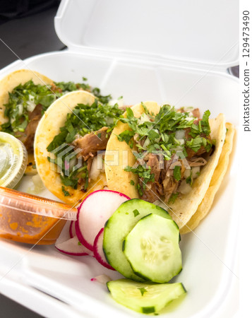 Three tacos topped with herbs and onions placed in a white takeout tray along with radish and cucumber slices. The image is bright and appetizing. Three tacos topped with herbs and onions placed in a white takeout tray along with radish and cucumber slices. The image is bright and appetizing. 129473490