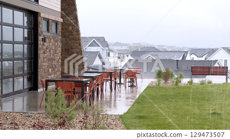 A stylish backyard with patio furniture and landscaped lawn is captured during a gentle rain. The stone building exterior and glass door frame the peaceful suburban scene with overcast light. A stylish backyard with patio furniture and landscaped lawn is captured during a gentle rain. The stone building exterior and glass door frame the peaceful suburban scene with overcast light. 129473507