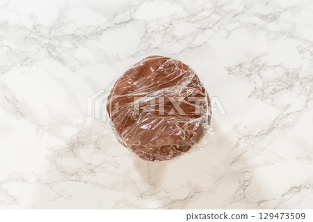 Close-up of pale gingerbread dough wrapped in plastic on a marble countertop for Pale Gingerbread Cookies with Subtle Spice Flavor. The dough is smooth and evenly shaped, ready for chilling. Close-up of pale gingerbread dough wrapped in plastic on a marble countertop for Pale Gingerbread Cookies with Subtle Spice Flavor. The dough is smooth and evenly shaped, ready for chilling. 129473509