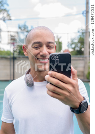 Latin fitness man influencer vlogging using smartphone on outdoor basketball court 129473531