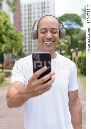 Latin bald man using phone while walking in city, digital lifestyle and active wellness concept 129473542
