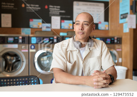 Bald Latin man sitting in laundromat with washing machines in background 129473552