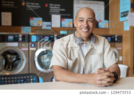 Bald Latin man sitting in laundromat with washing machines in background 129473553