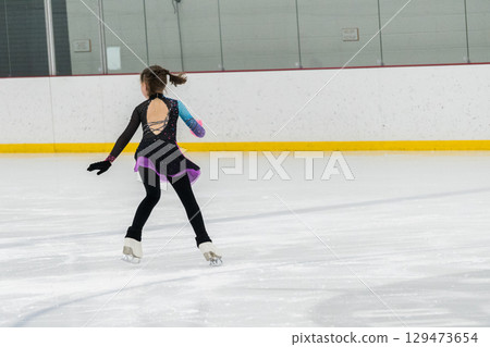 Young girl perfecting her figure skating routine while wearing her competition dress at an indoor ice rink. 129473654