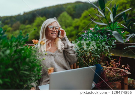 Mature woman sitting on balcony making phone call, working remotely. 129474896
