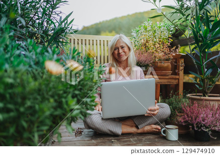 Older woman working from homeoffice, sitting on terrace with laptop. 129474910