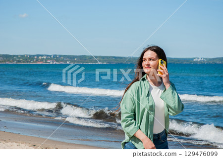 Smiling young woman with long brown hair strolling by the sea shore and talking on a yellow smartphone, enjoying a sunny day and relaxed coastal atmosphere 129476099
