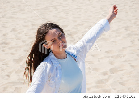 Young woman with long brown hair standing on sandy beach with eyes closed and arms raised, enjoying the sunlight, symbolizing freedom, happiness, and summer vacation Young woman with long brown hair standing on sandy beach with eyes closed and arms raised, enjoying the sunlight, symbolizing freedom, happiness, and summer vacation 129476102