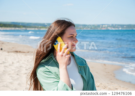 Smiling woman with long hair talking on a yellow smartphone while standing on a sandy beach near the sea waves under a sunny blue sky enjoying summer freedom and digital nomad lifestyle 129476106