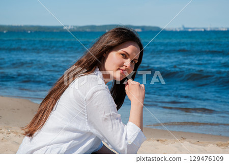 Young woman with long brown hair sitting on sandy beach near the sea on a sunny day, looking at the camera, symbolizing summer vacation, travel, and coastal lifestyle Young woman with long brown hair sitting on sandy beach near the sea on a sunny day, looking at the camera, symbolizing summer vacation, travel, and coastal lifestyle 129476109