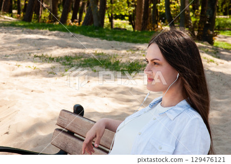 Young woman with long brown hair sitting on a wooden bench in a sunny sandy park, listening to music with earphones and enjoying a peaceful moment, symbolizing mindfulness and digital lifestyle 129476121