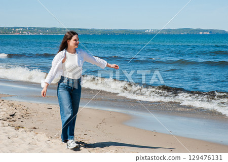 Young woman with long brown hair walking along sandy beach near the sea on a sunny day, smiling and enjoying summer vacation, symbolizing freedom, travel, and coastal lifestyle 129476131