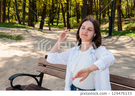 Young woman with long brown hair sitting on a wooden bench in a sunny sandy park, enjoying music with earphones and dancing with eyes closed, symbolizing happiness, freedom, and mindful lifestyle 129476133