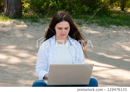 Young woman with long brown hair sitting on sandy ground in a sunny park, working on a laptop, symbolizing remote work, freelancing, and digital nomad lifestyle Young woman with long brown hair sitting on sandy ground in a sunny park, working on a laptop, symbolizing remote work, freelancing, and digital nomad lifestyle 129476135