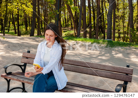 Young woman with long brown hair sitting on a wooden bench in a sunny sandy park, using a smartphone and enjoying a relaxing moment in nature, symbolizing digital lifestyle and remote work outdoors Young woman with long brown hair sitting on a wooden bench in a sunny sandy park, using a smartphone and enjoying a relaxing moment in nature, symbolizing digital lifestyle and remote work outdoors 129476147