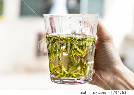 Person holding transparent glass with longjing tea infused in hot water to observe the quality. 129476301