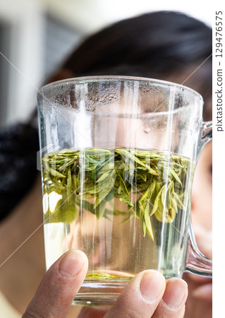 Person holding transparent glass with longjing tea infused in hot water to observe the quality. 129476575