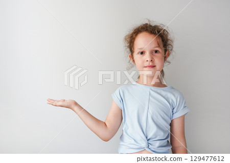 A child girl stands against a plain background, holding one hand out with an open palm as if presenting something A child girl stands against a plain background, holding one hand out with an open palm as if presenting something 129477612