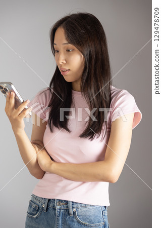 A young woman in a pink t-shirt and jeans is looking at her smartphone with a focused expression 129478709