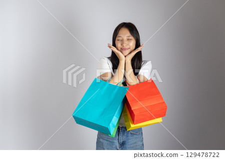 Young woman is holding several shopping bags and smiling joyfully with closed eyes, placing her hands on her cheeks 129478722