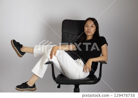Young woman is sitting casually on an office chair with her legs crossed and resting on another part of the chair, looking Young woman is sitting casually on an office chair with her legs crossed and resting on another part of the chair, looking 129478738