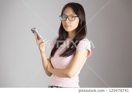 A young woman wearing glasses and a pink t-shirt, holding a silver mobile phone in her right hand while looking directly at the  129478741