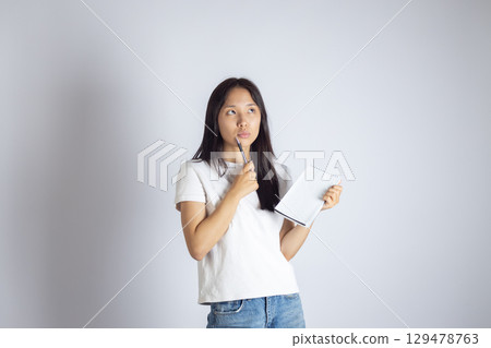 Young asian woman is shown contemplating while holding a pen to her lips and a notebook in her other hand. Young asian woman is shown contemplating while holding a pen to her lips and a notebook in her other hand. 129478763