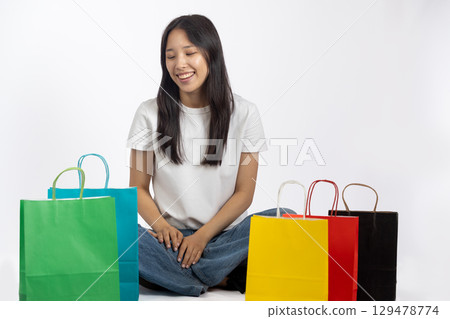 A young woman is sitting cross-legged on the floor, smiling and looking down at several colorful shopping bags placed around  129478774