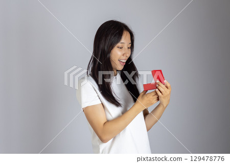 Woman in white t shirt is smiling with a surprised expression as she opens a small red gift box 129478776