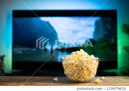 A glass bowl of popcorn and remote control in the background the TV works. Evening cozy watching a movie or TV series at home 129479616